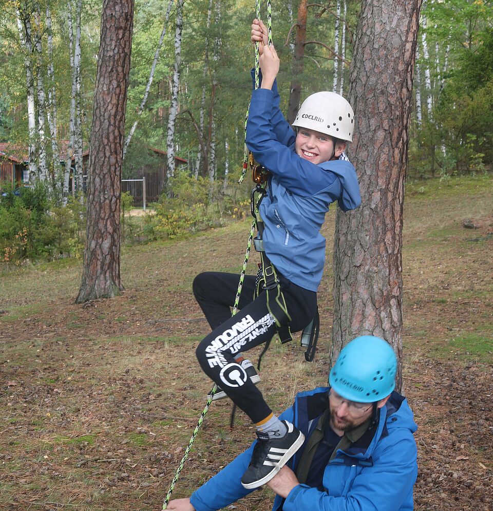 Junge mit Klettergurt und Helm hängt an Seil und schaut in Kamera