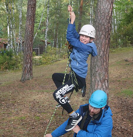 Junge mit Klettergurt und Helm hängt an Seil und schaut in Kamera